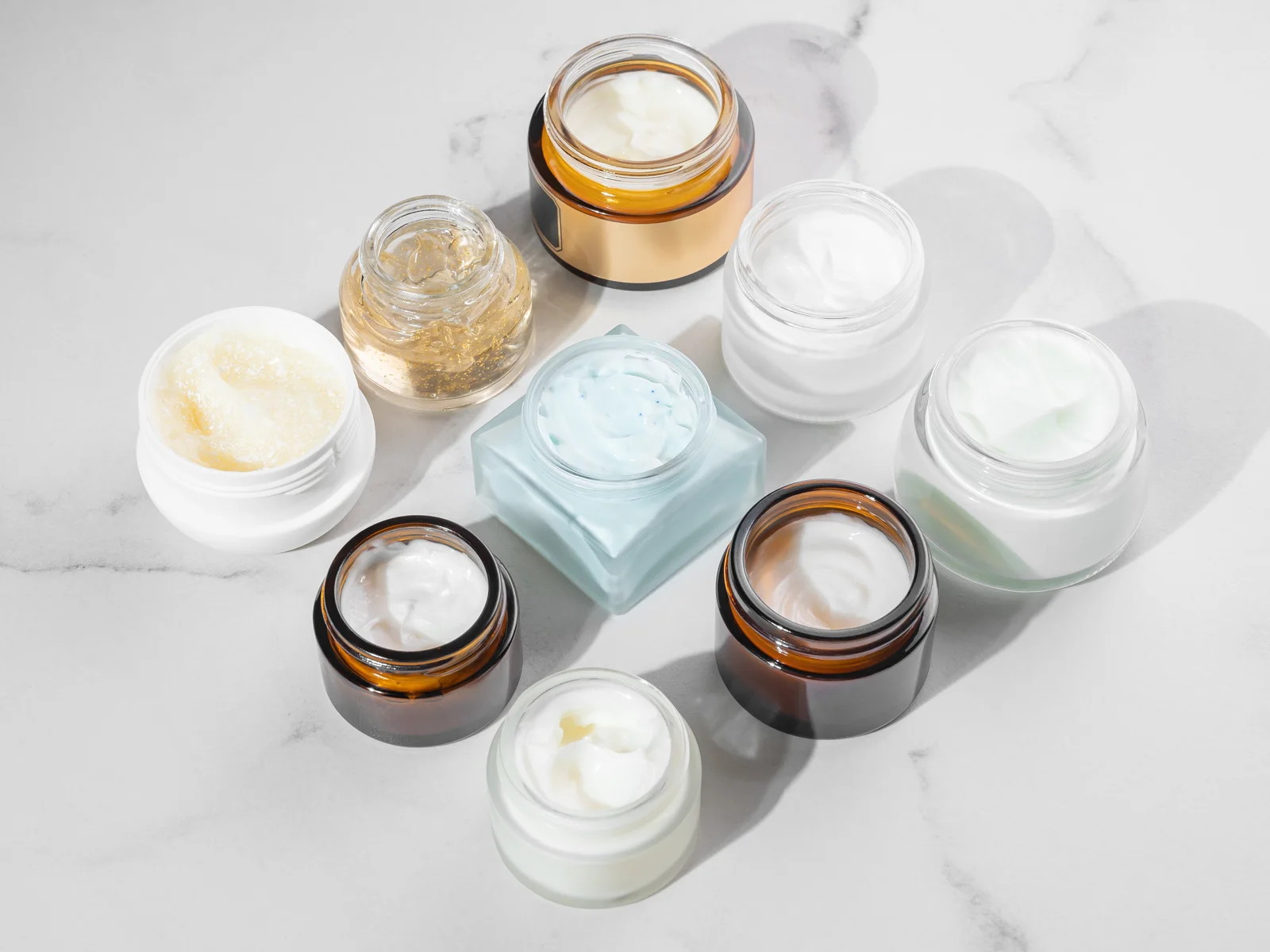 Assorted open jars of skincare creams and gels on a marble surface